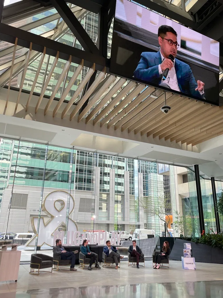 Five people sit in chairs with mics in their hands on stage at The Ampersand in Calgary, Alberta during Plug and Play's Sustainability panel discussion. A screen above projects the speaker, CEO of Pani, to the audience.
