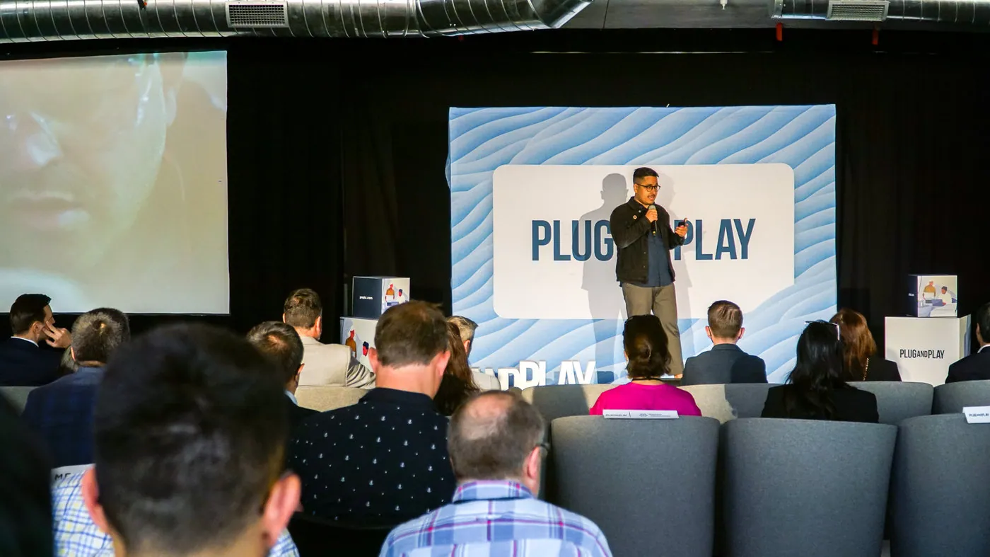 CEO of Pani on stage with a screen next to him showing a slide that describes three case studies that the platform has been used in. A Plug and Play sign is behind the speaker.