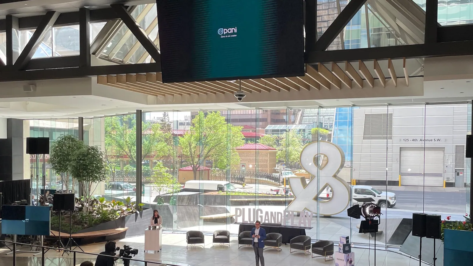 A large screen displaying the Pani logo hangs above the Plug and Play lobby in Calgary, with the Plug and Play sign visible through the glass wall behind two speakers