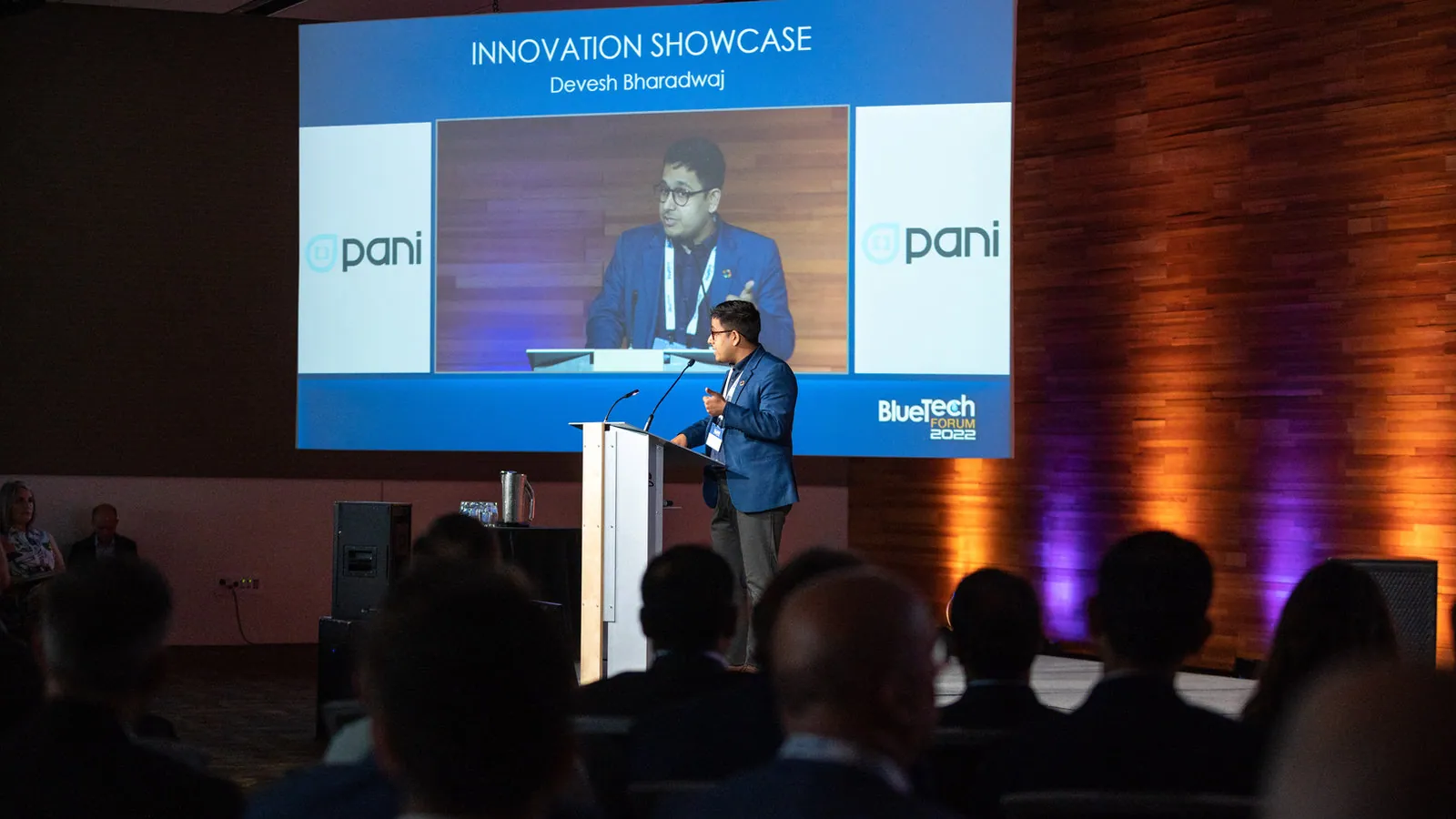 A crowd watches a person speaking on stage in front of a projected screen reading 'Innovation Showcase, Devesh Bharadwaj, Pani, BlueTech Forum 2022'